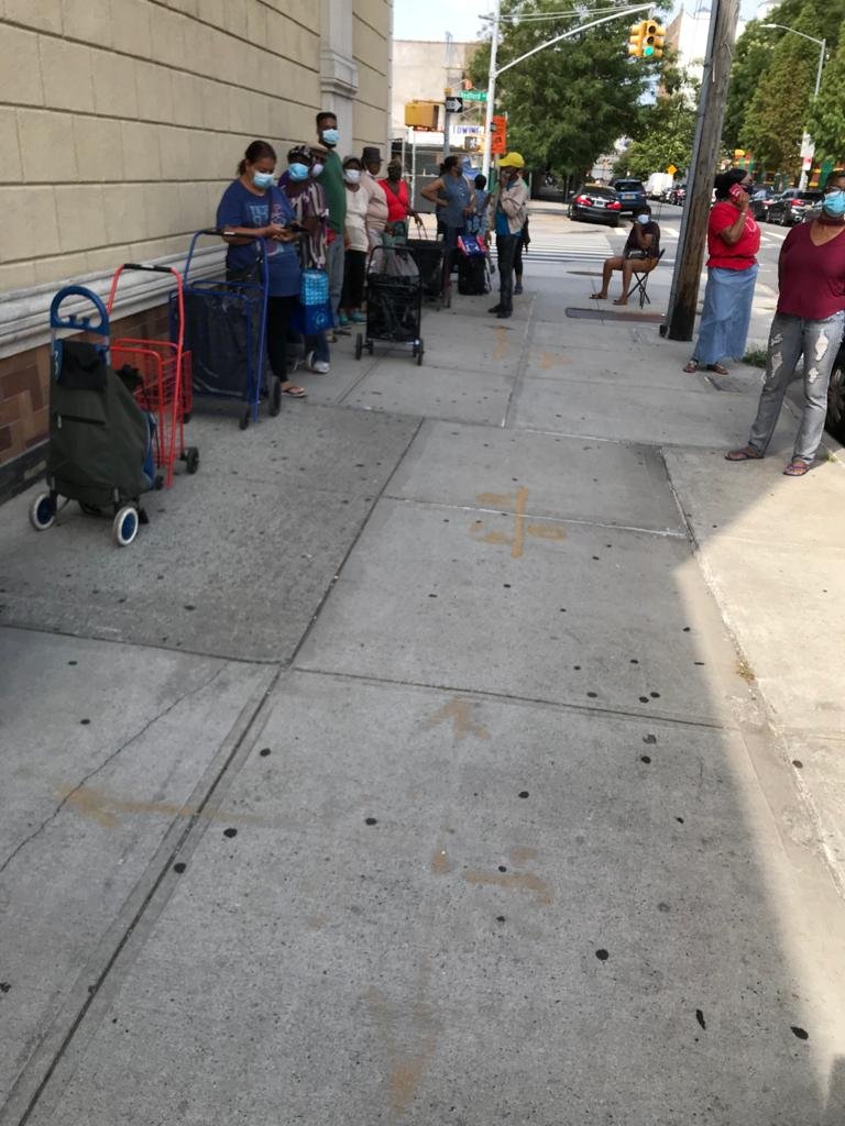 People standing in line outside on the sidewalk wearing masks, waiting for service.