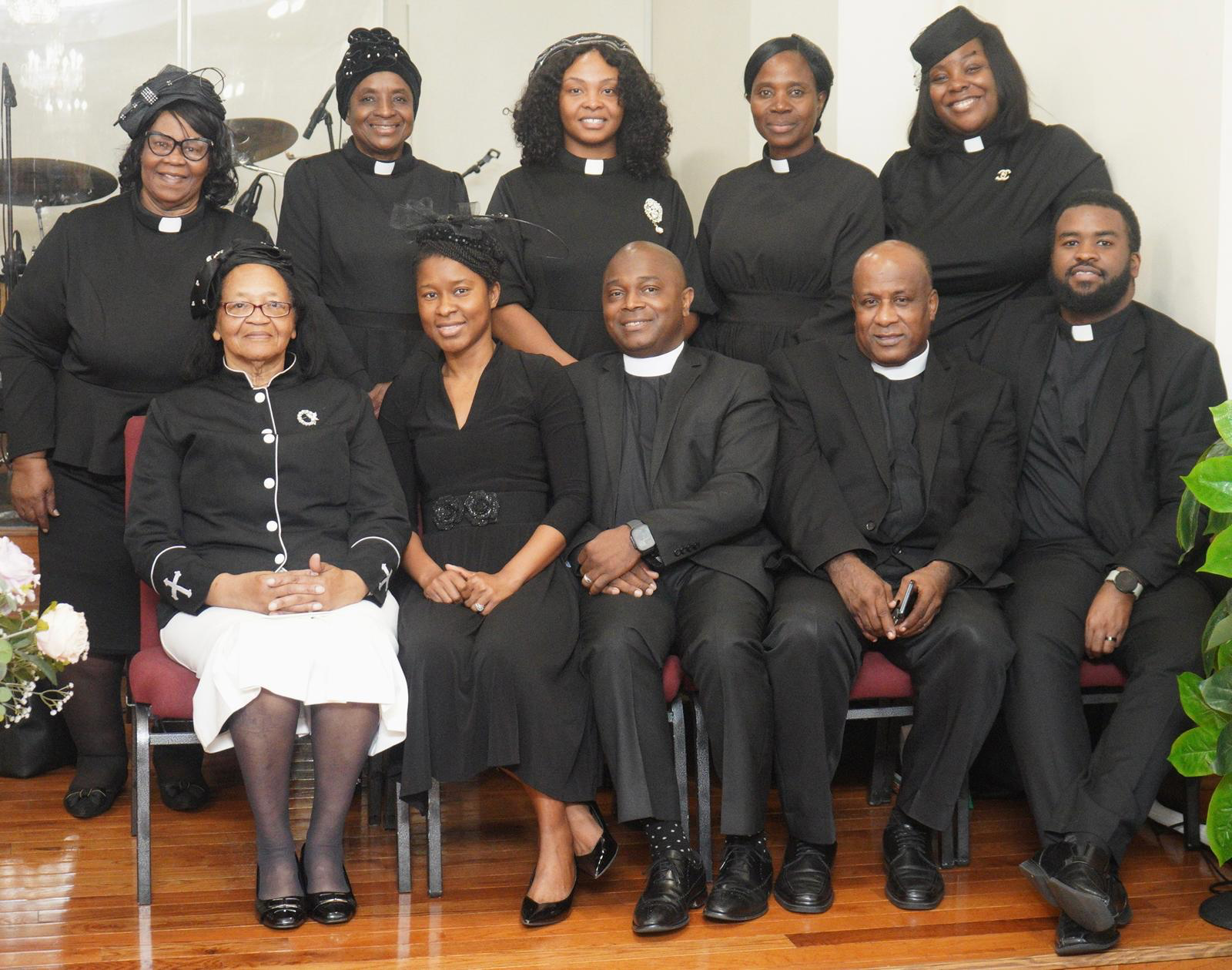 A group of eleven people, mainly women and some men, dressed in black religious attire with collar insignias, posed together indoors, sitting and standing, with microphones and musical instruments visible in the background.