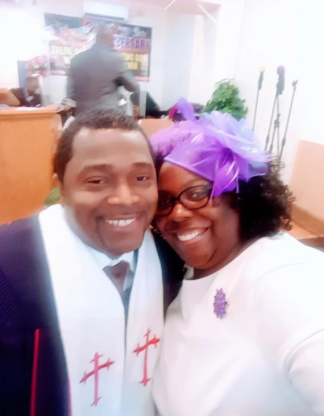 Two smiling people taking a selfie at a church event, the man is wearing a white robe with red crosses and the woman is wearing glasses, a purple head wrap, and a white outfit with a purple pin.
