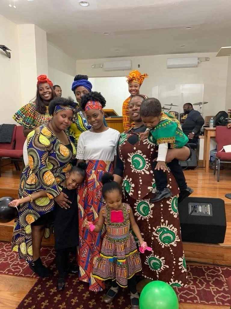 A group of African women, children, and a man in a room celebrating, wearing colorful traditional African clothing and headwraps. They are standing on a wooden floor with balloons around.