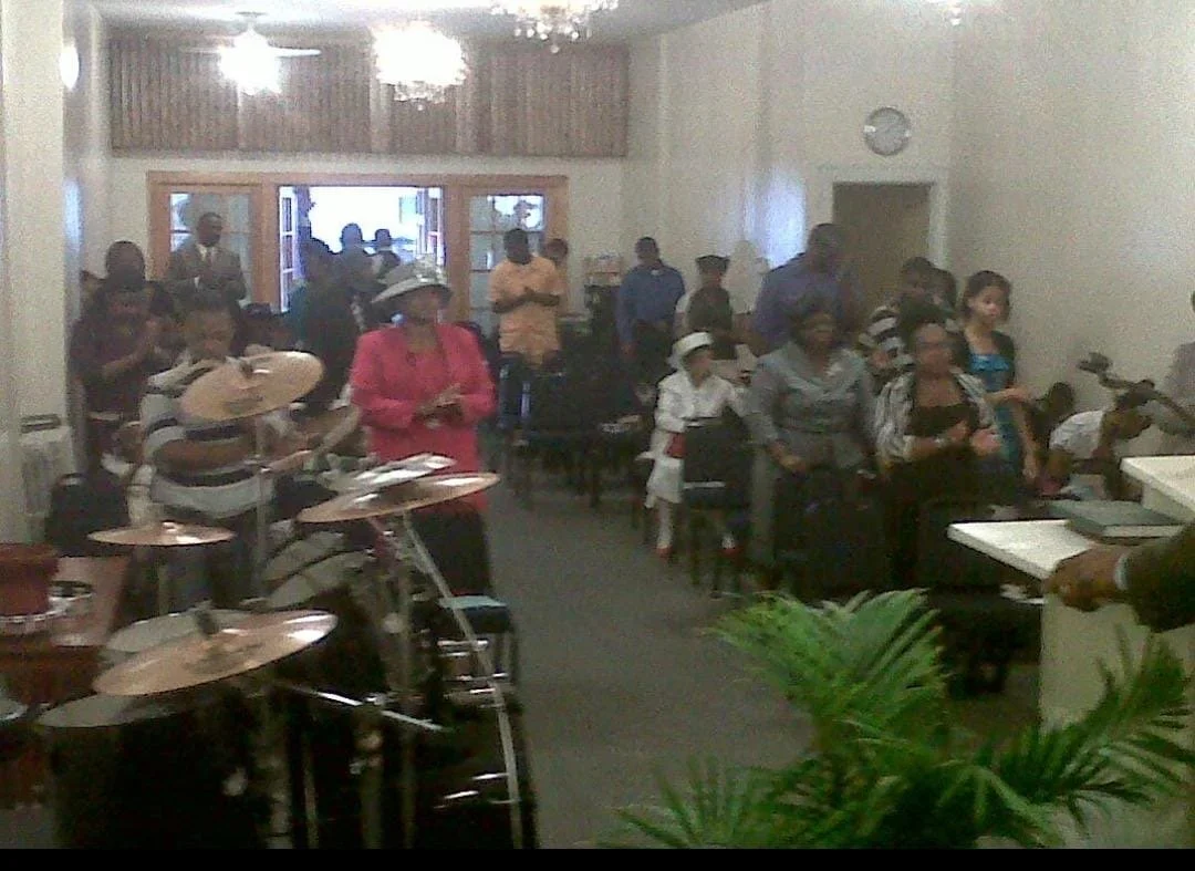 People standing and sitting inside a church during a service or event, with musical instruments and a large plant in the foreground.