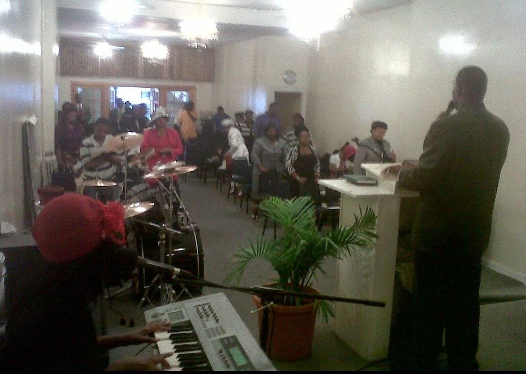 A church service with a congregation of people sitting in pews and chairs, a person playing a keyboard, and a drummer in the front near the pulpit, with the pastor speaking to the congregation.