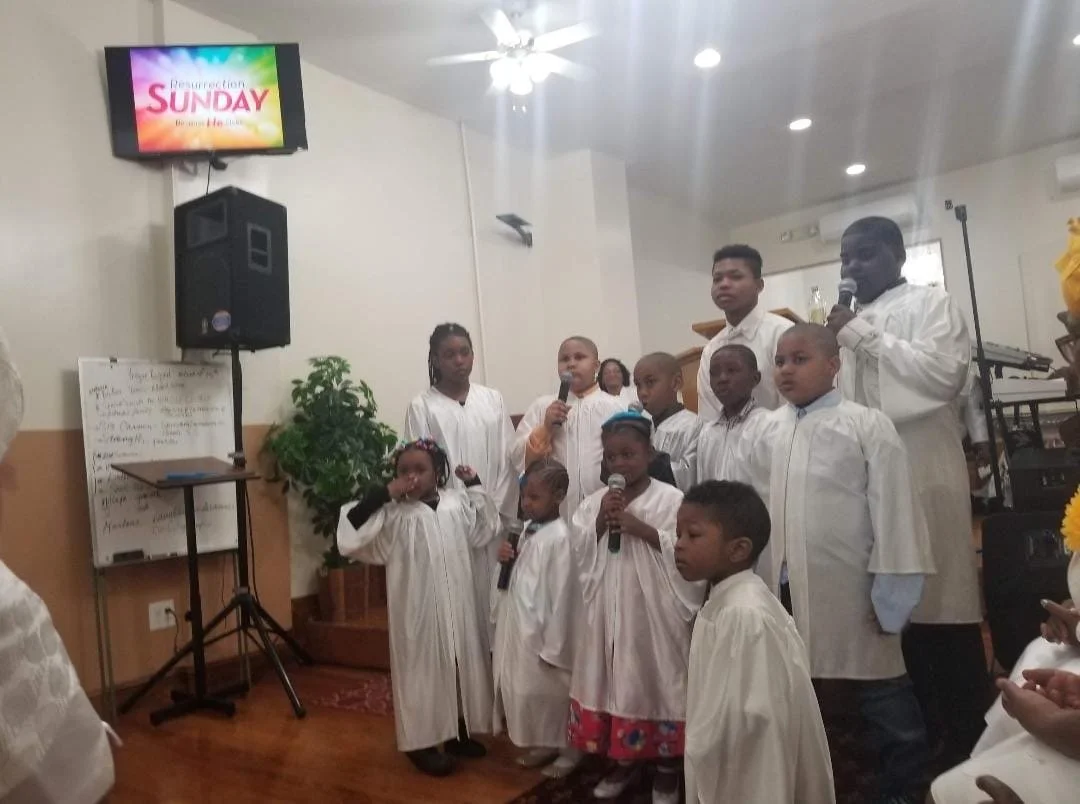 Children and adults in white robes singing or speaking into microphones during church service inside a chapel with a speaker, a plant, a whiteboard, and a digital screen displaying "Resurrection SUNDAY."