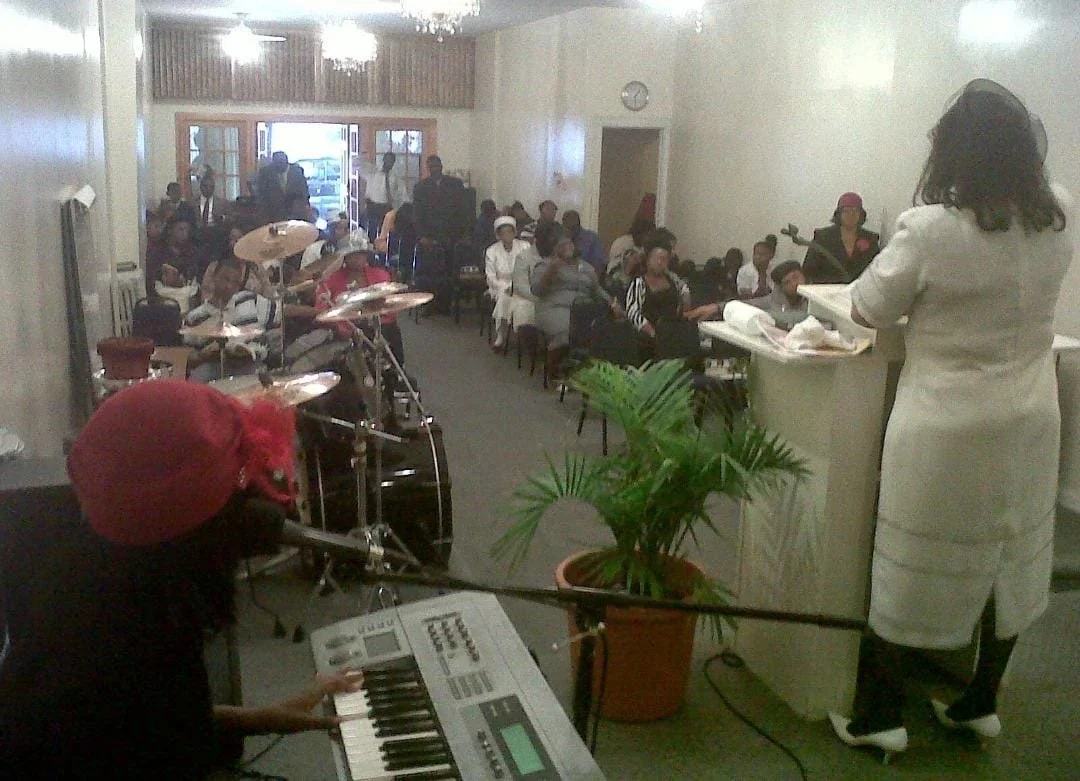 A woman on stage playing a keyboard at a community event, with a choir seated behind her and a drummer nearby. An audience is sitting and standing, watching, in a room decorated with chandeliers, a potted plant, and a clock on the wall.
