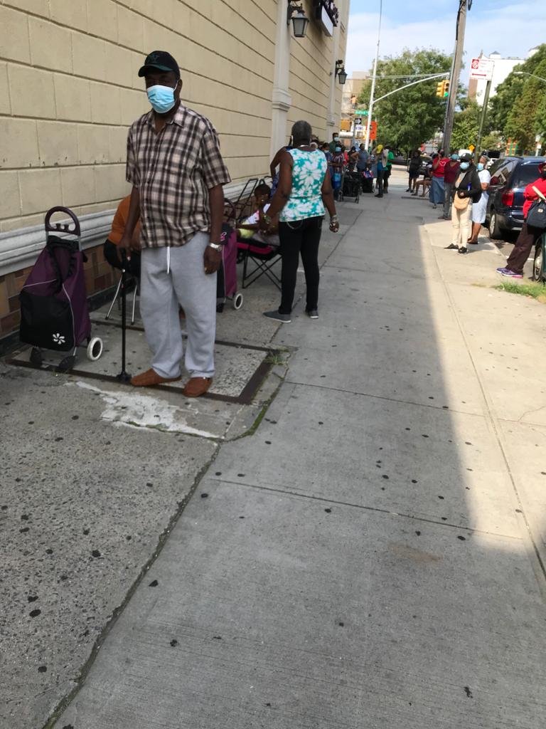 A man wearing a face mask, plaid shirt, and gray pants standing on a city sidewalk next to a shopping cart with a purple bag, with several other people in line or waiting nearby on the sidewalk, with cars parked along the street and traffic lights in