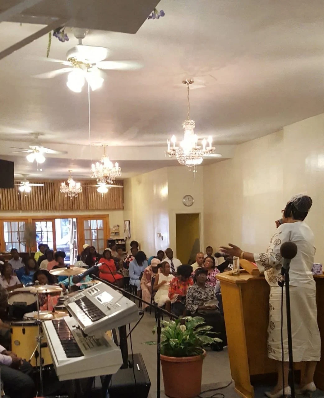 A woman in white attire speaking at a podium in a church or community hall, with an audience seated facing her, musical instruments including a keyboard and drums in the foreground, chandeliers and ceiling fans overhead, wooden paneling and windows i