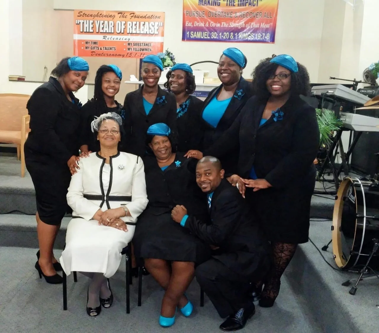Group of nine people, mostly women, dressed in black with blue accents, some wearing blue head wraps, gathered in a church or event setting, with musical instruments and church banners in the background.