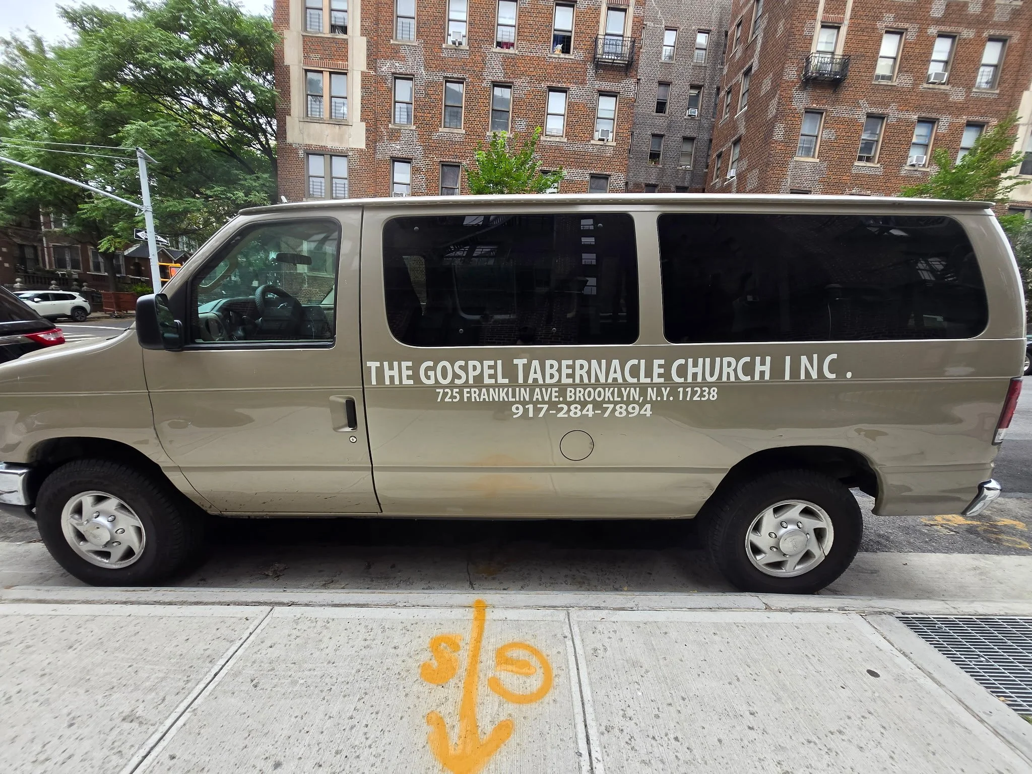 A beige van parked in a parking lot with text advertising The Gospel Tabernacle Church Inc., located at 725 Franklin Ave, Brooklyn, NY, with a phone number 917-284-7894. The parking lot has yellow spray-painted markings and is adjacent to a brick residential building with multiple windows and some small balconies. There are green trees and other parked cars visible.