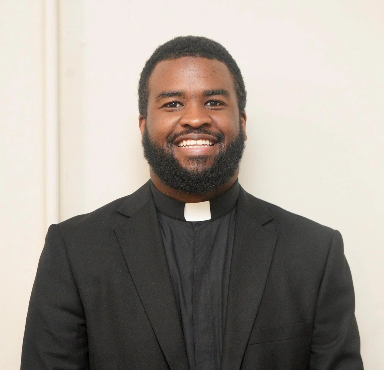 Smiling man in black clergy suit with a white clerical collar standing in front of a plain white wall.