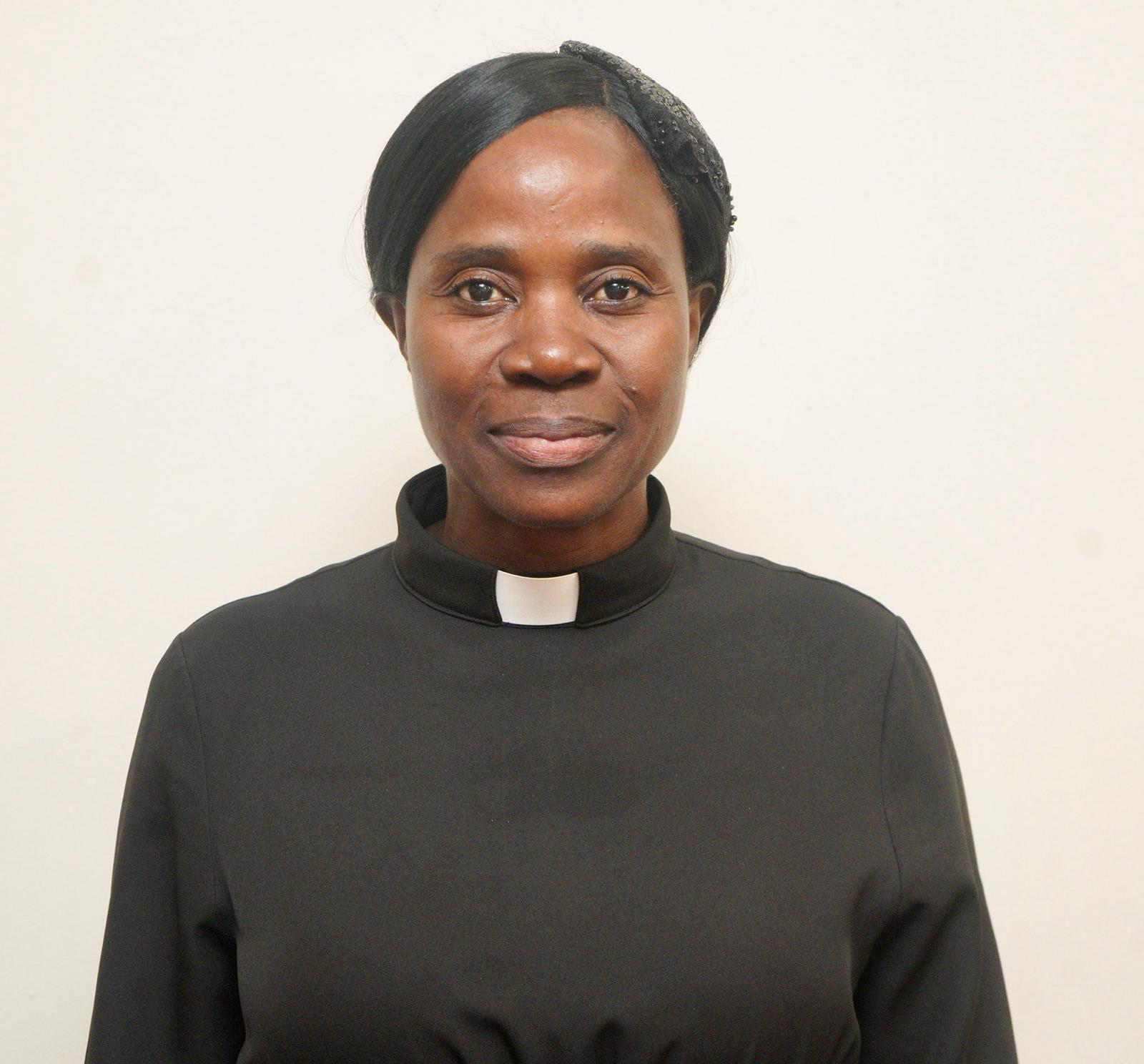A woman dressed in a black clerical shirt with a white collar, standing in front of a plain white background.