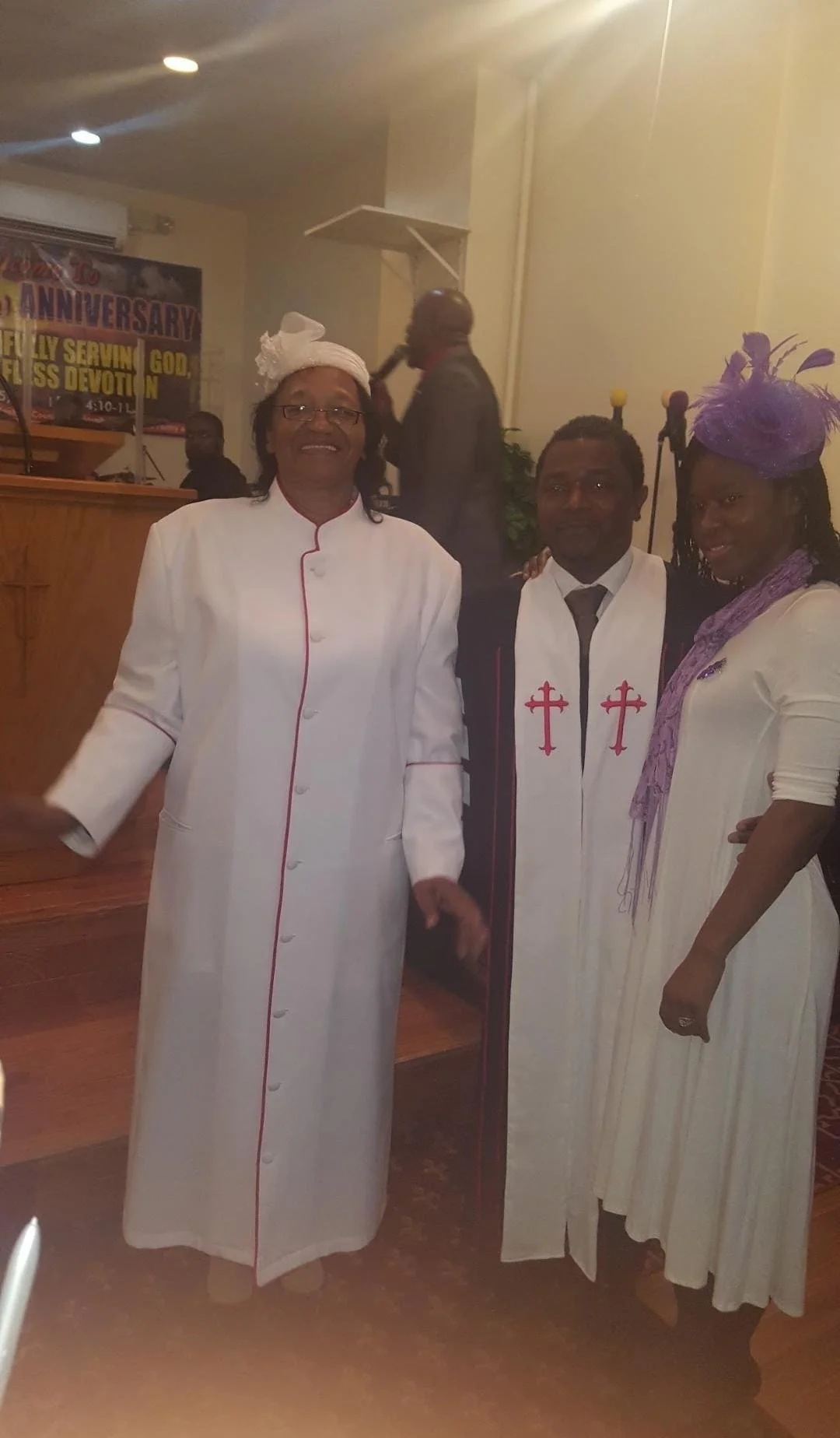 Three people wearing religious robes and hats, standing inside a church. Two men are dressed in white robes with red and purple crosses, and a woman is dressed in a white dress with purple accessories. They are posing together, smiling at the camera.