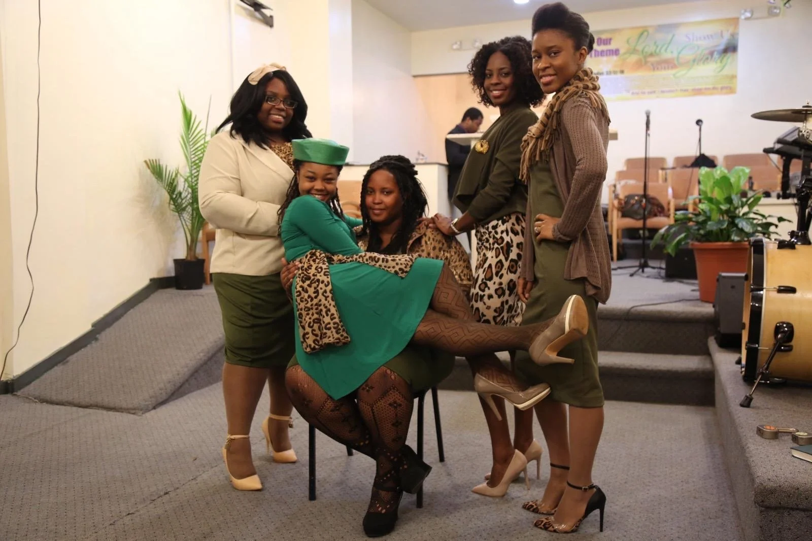 Group of five women celebrating on stage, one woman sitting on a chair with her legs lifted by others, all dressed in green and animal print clothing, in a church or community hall setting.