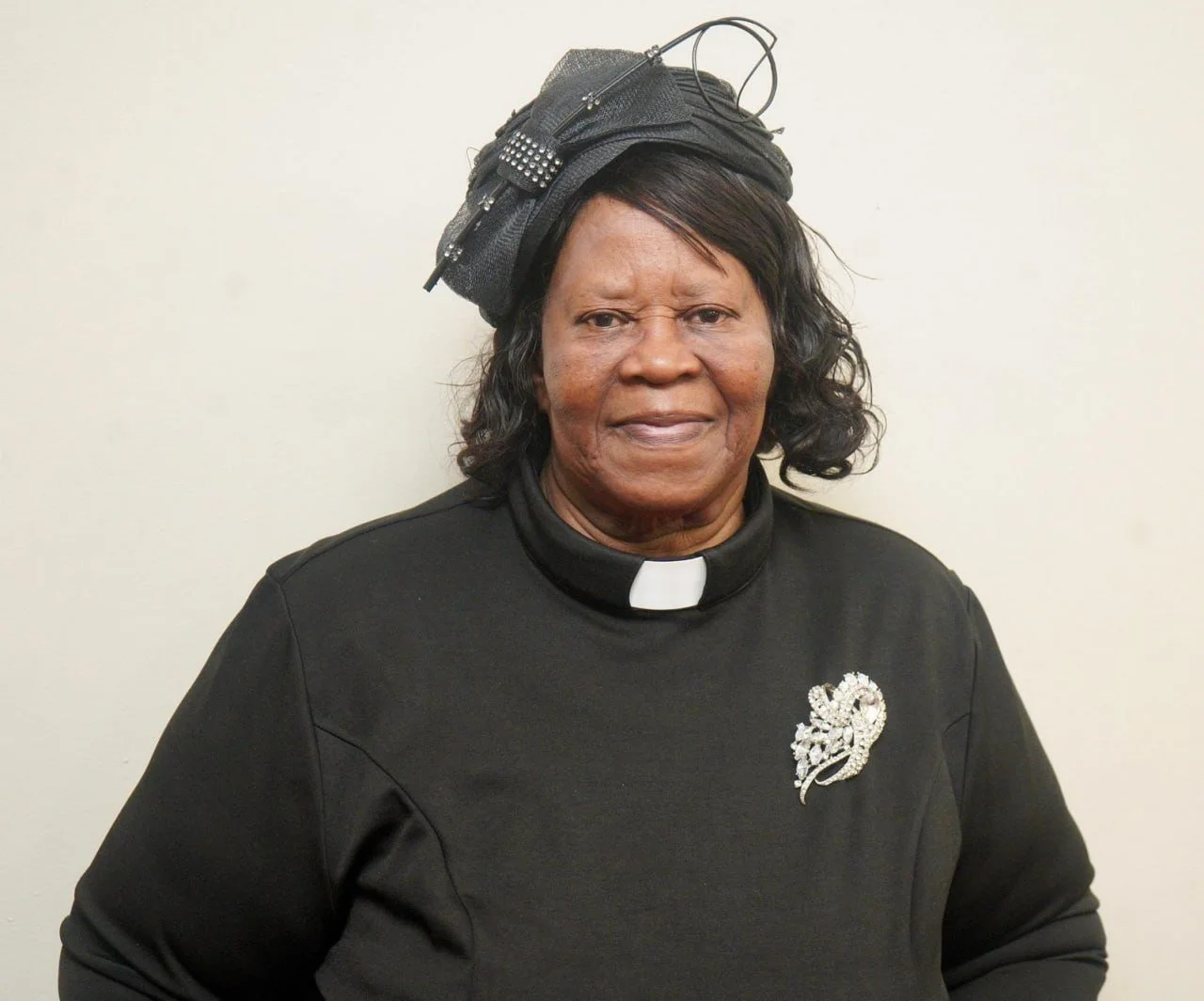 An elderly woman wearing a black outfit with a white clerical collar and a decorative brooch, standing against a plain background. She has a black hat with fabric and embellishments, and she has a serious expression.