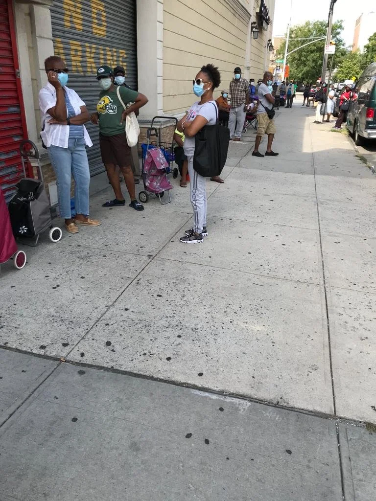 People wearing masks standing on a city sidewalk, some in line, near a building with a closed storefront.