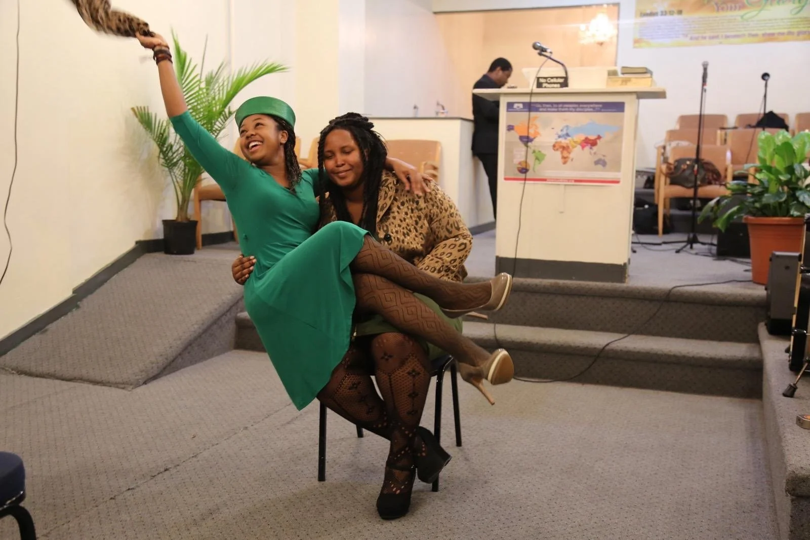 Two women are sitting on a chair in a room, one dressed in vibrant green, holding a phone, taking a selfie, with both smiling. The other woman, wearing animal print and patterned tights, sits smiling with her arm around the woman in green. The backgr