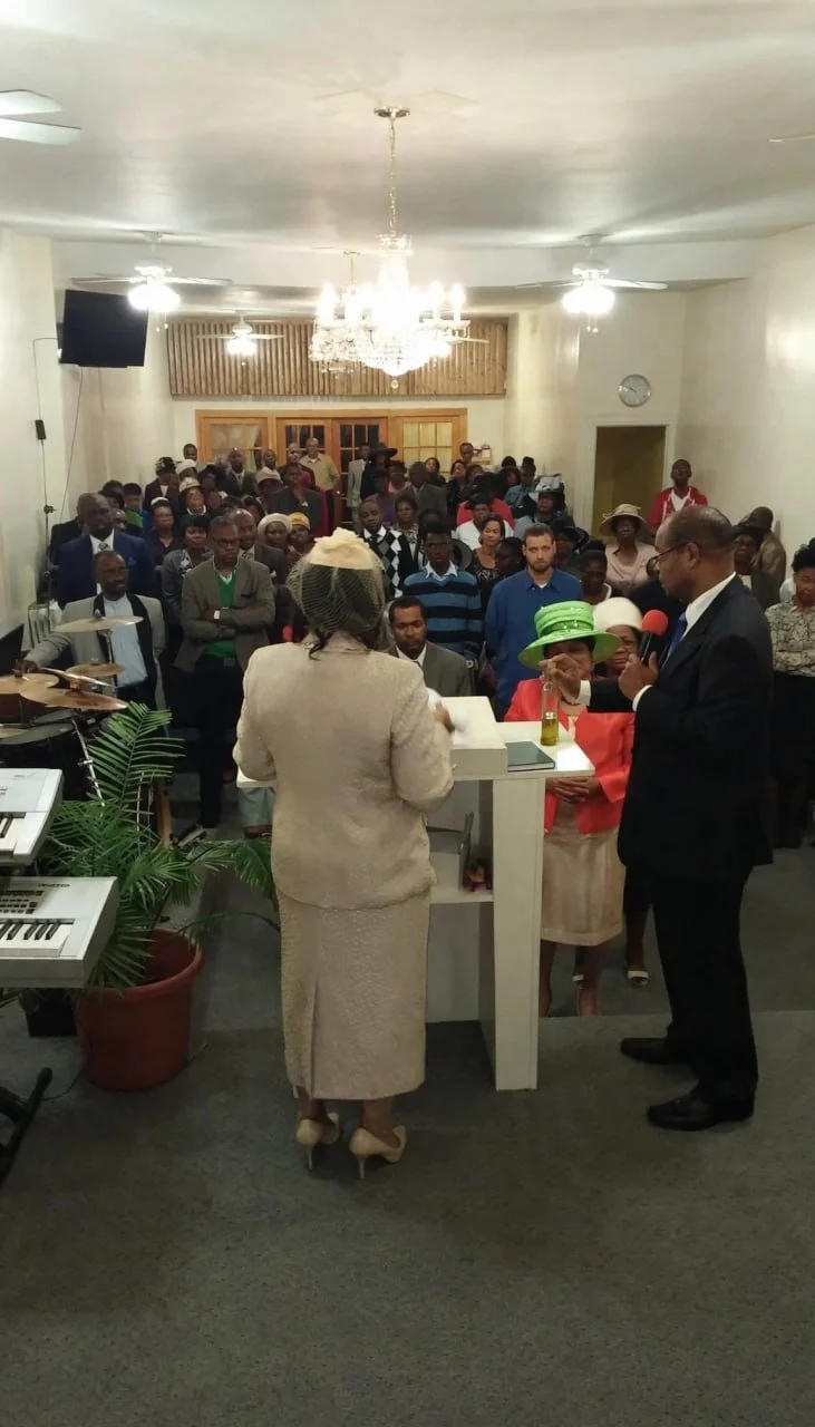 A religious service or ceremony with a woman and man at the podium, and an audience in the background. The woman is dressed in a light-colored outfit and a hat, and the man is in a dark suit with glasses and a microphone. The room has a chandelier, c