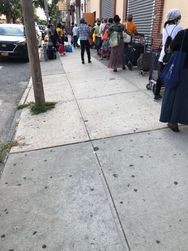 People standing in line outside a store on a city sidewalk, some with shopping carts and bags, with cars parked along the street.