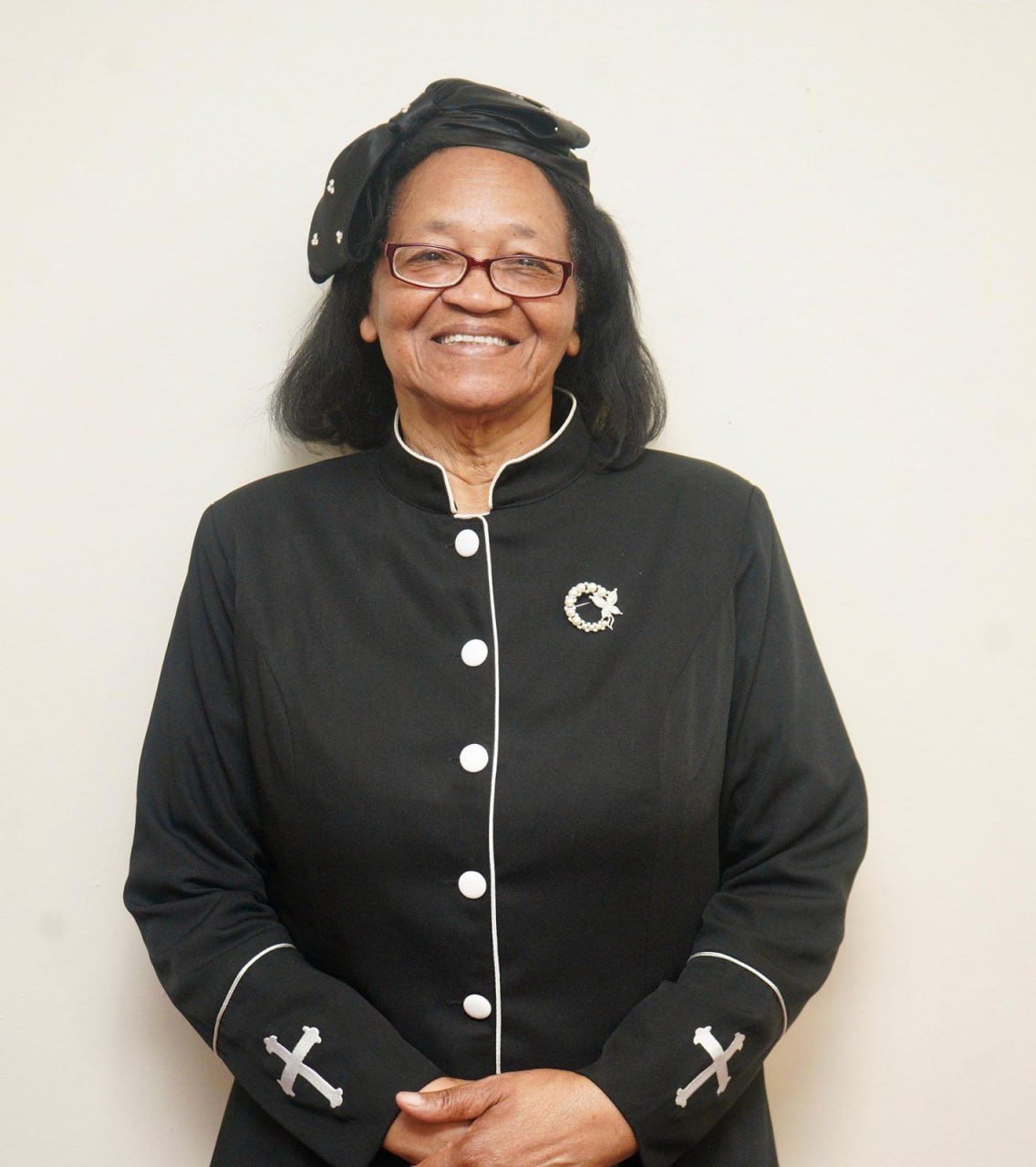 An elderly woman with glasses, wearing a black dress with white trim, a brooch, and a headscarf, standing and smiling against a plain background.