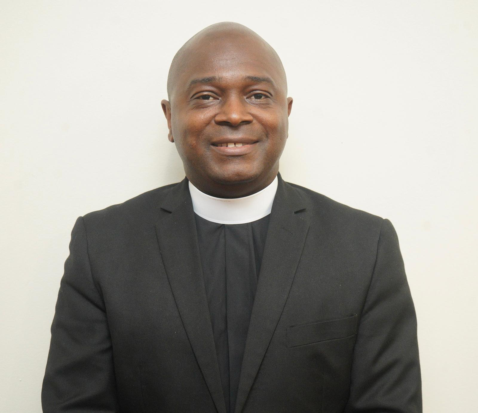 A man dressed in a black clerical suit with a white clerical collar, smiling, against a plain, light-colored background.