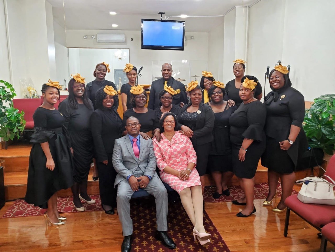Group of women in black dresses with yellow headbands, two people in the front with one man in gray suit and a woman in pink dress seated, inside a room with wood flooring and plants.