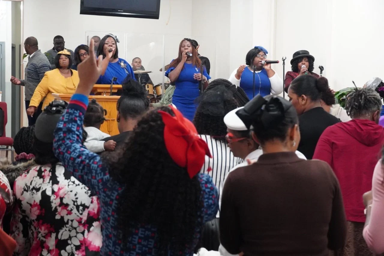 A group of women singing and performing on stage with a crowd of people watching, some raising their hands, in an indoor setting.