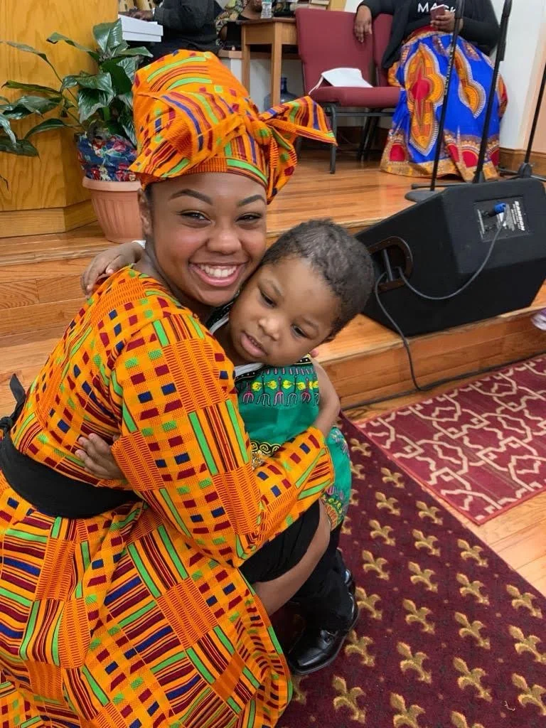 A woman and a young boy hugging. The woman is wearing a colorful traditional African dress and headwrap, smiling broadly. The boy is dressed in a green shirt with colorful embroidery, looking off to the side. They are inside a room with wooden floors
