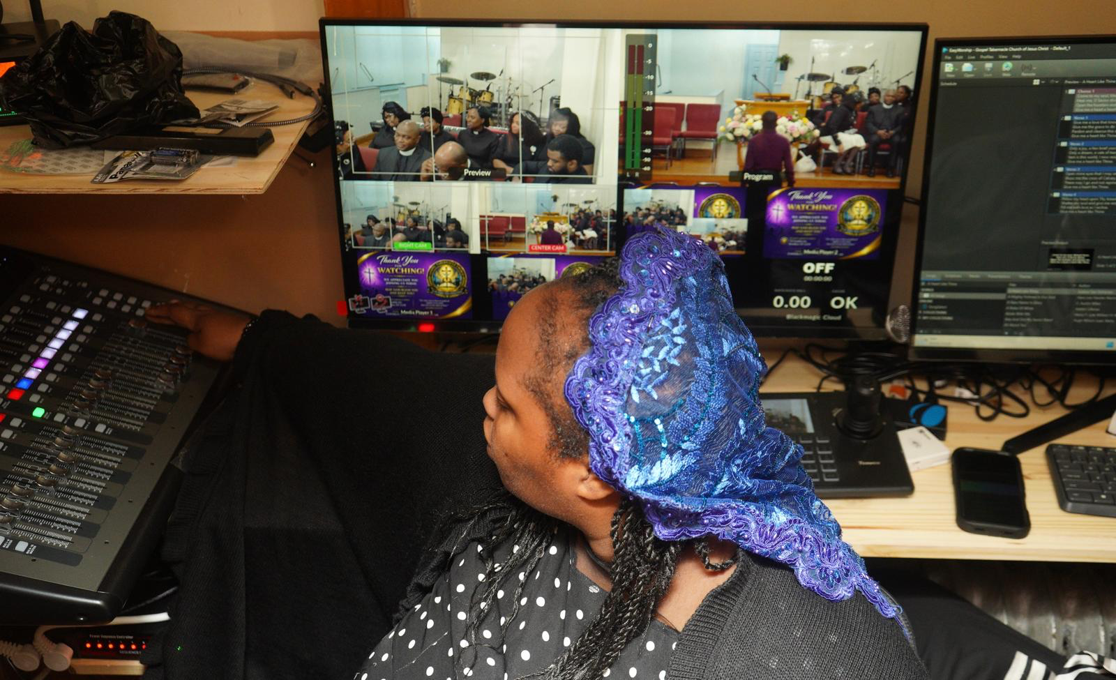 A person wearing a dark top and a purple lace headscarf working at a video control station with a large monitor displaying a church service with attendees.