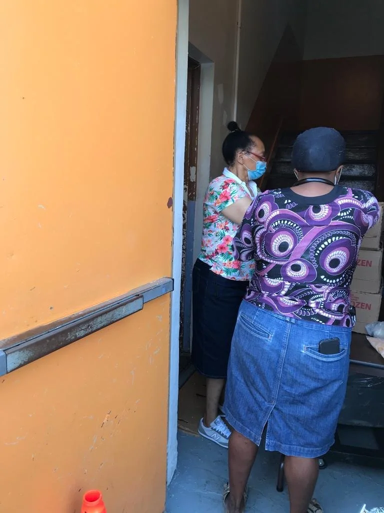 Two people, a woman wearing a face mask, glasses, floral shirt, and skirt, and a man in a patterned shirt and denim skirt, are standing inside a doorway surrounded by boxes.