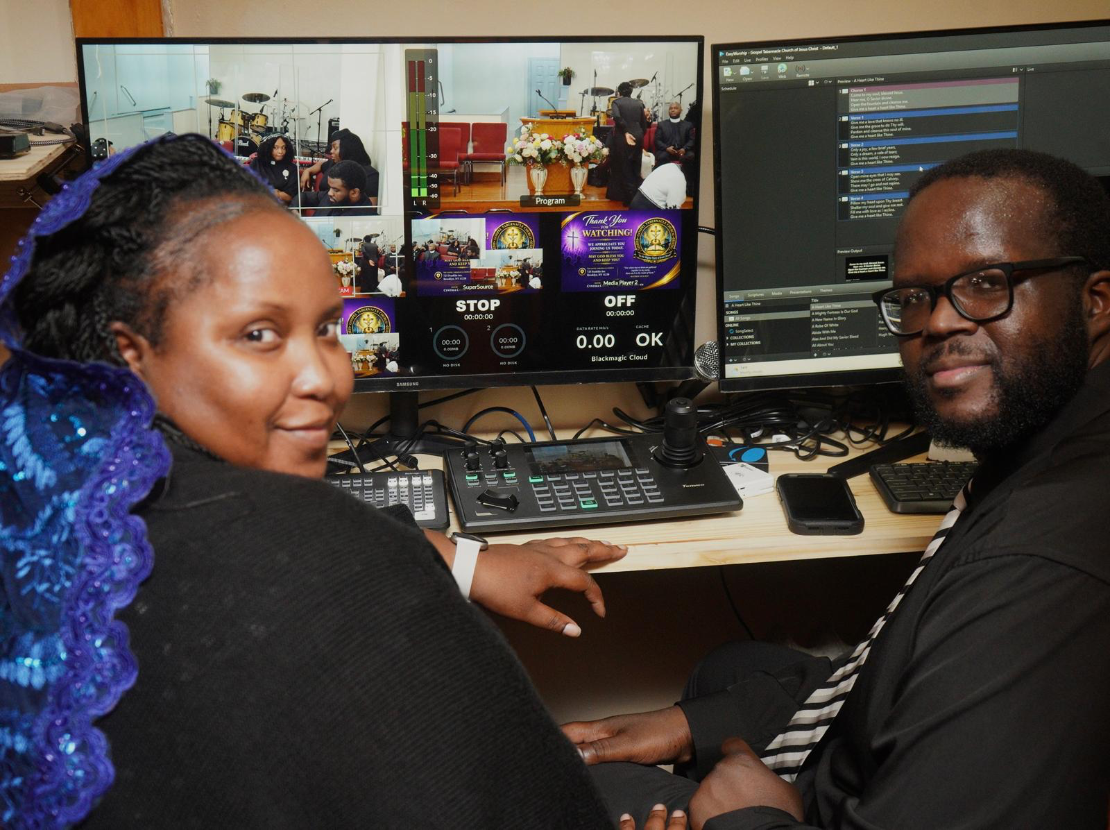 Two individuals sitting in front of a video production control station with multiple monitors, audio equipment, and a computer, inside a church setting.