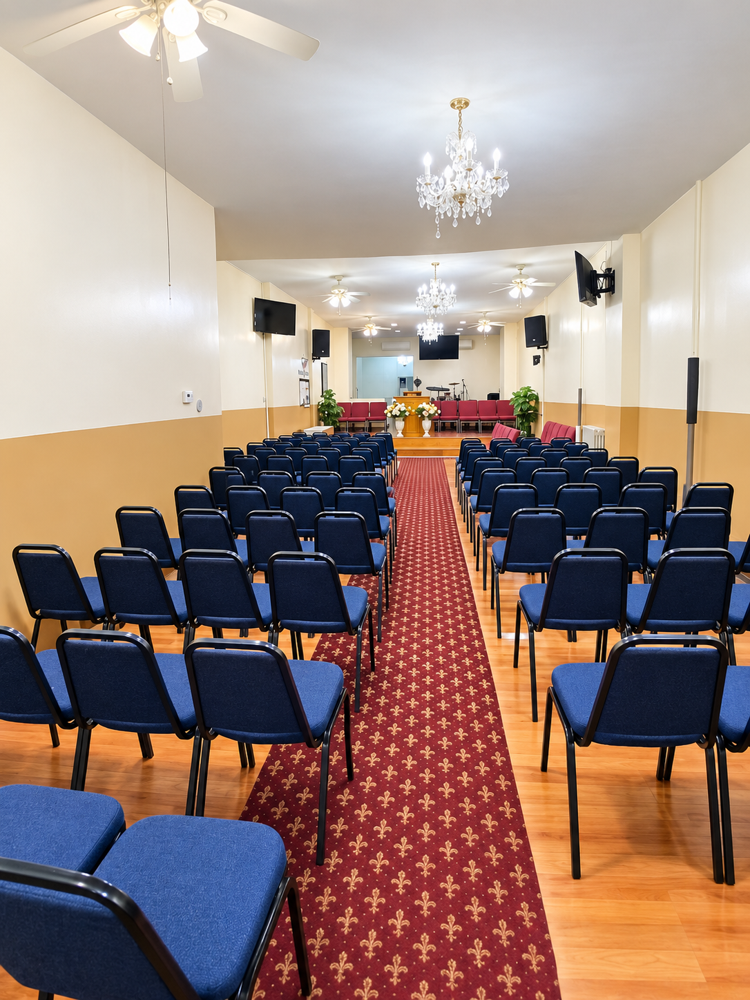 Empty church or event hall with rows of blue chairs, red carpet aisle, stage with flowers, chandeliers, ceiling fans, and mounted screens.