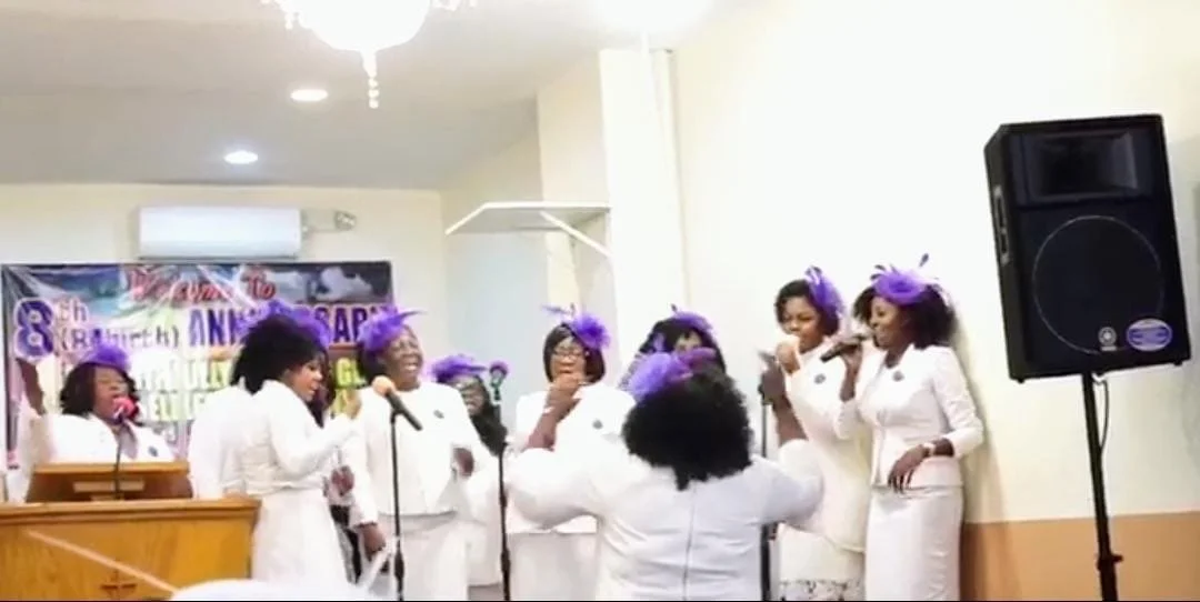 A group of women dressed in white, wearing purple headpieces, are singing or performing on stage at an indoor event, with a woman facing them in the foreground.