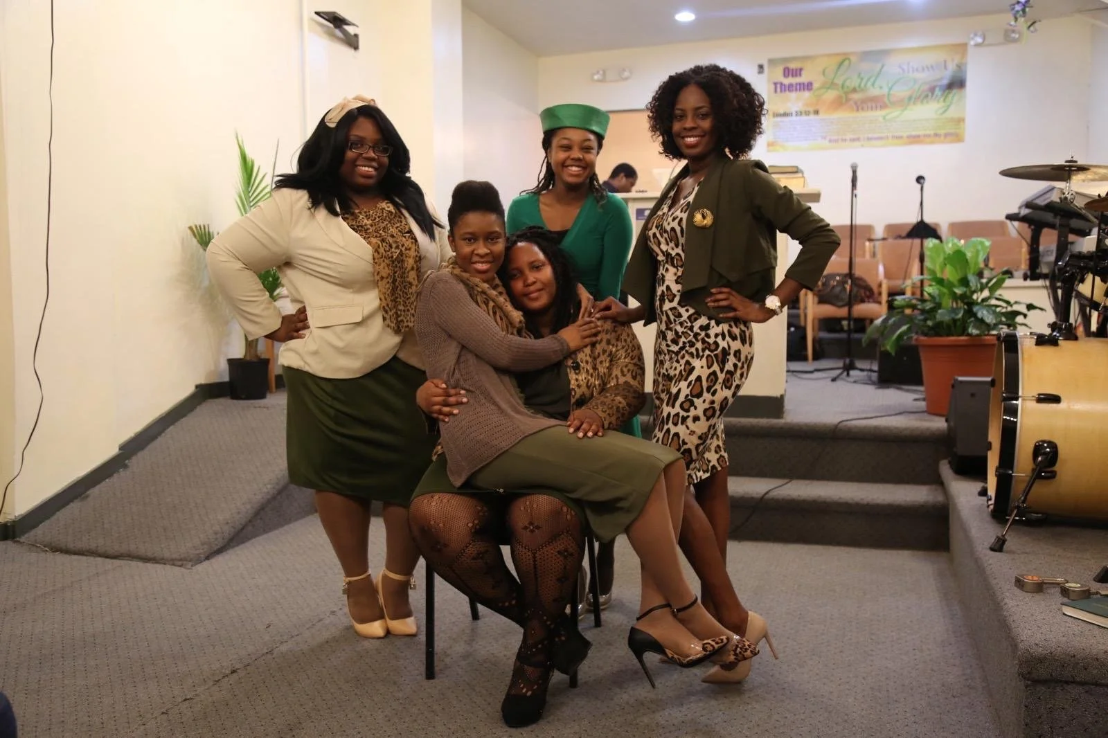 Group of six women posing and smiling inside a church or event hall with musical instruments and a banner in the background.
