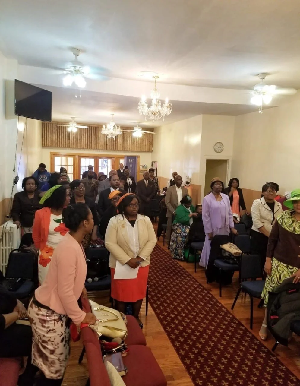 People standing and sitting in a room with chandeliers, ceiling fans, and a clock on the wall, gathered for an event or meeting.