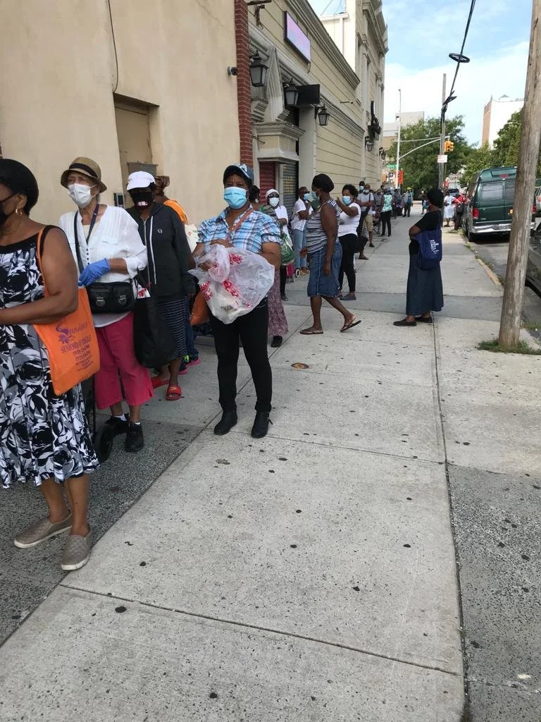 People standing in a long line outside on a city sidewalk, all wearing masks, some with umbrellas or bags, waiting to enter a building.