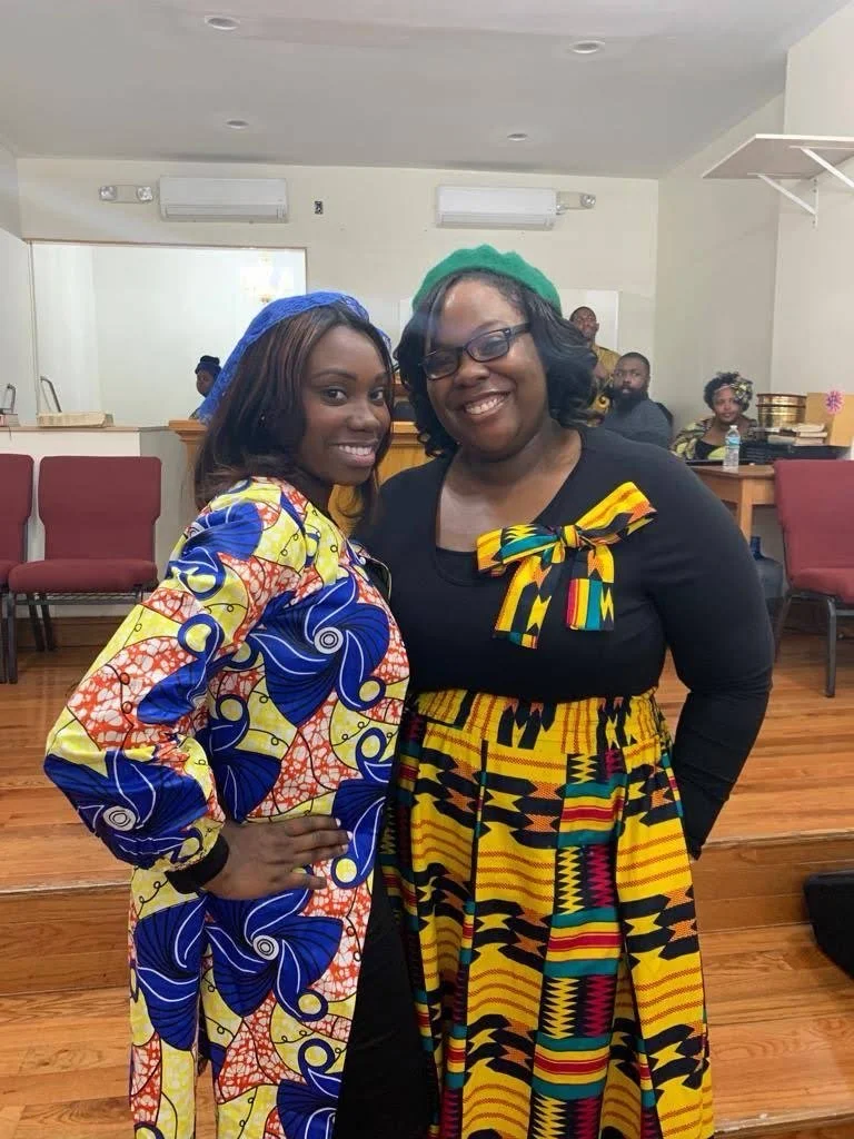 Two women smiling and standing close together, dressed in colorful traditional African clothing, in an indoor setting with chairs and people in the background.