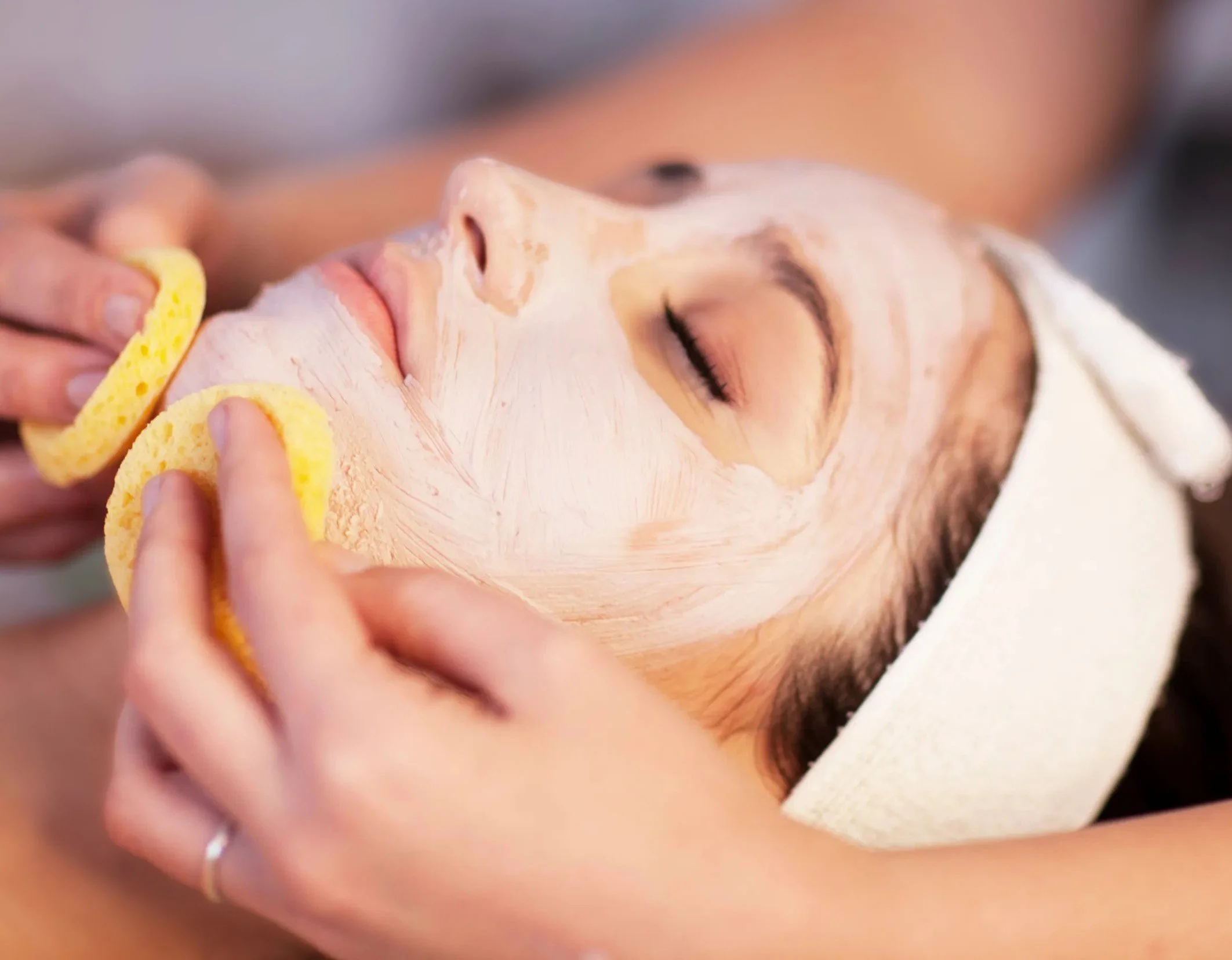 A person is relaxing with their eyes closed while applying a facial mask. A sponge is being used to apply the mask to their face, and a white towel is draped over their head.