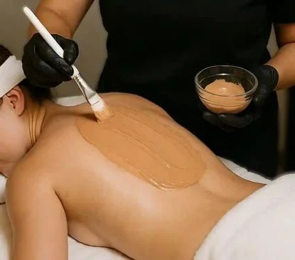 Person receiving a back massage with a massage brush, while another person holds a bowl of cream or lotion.