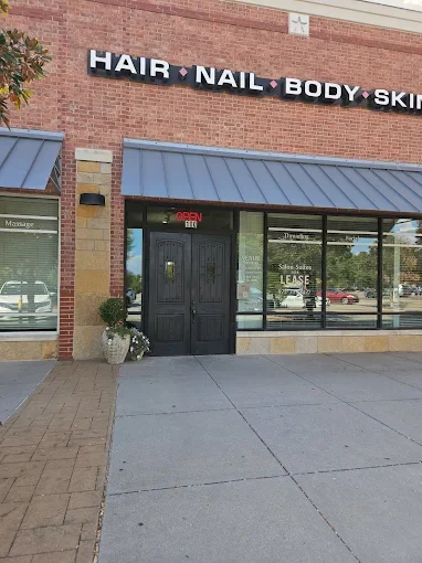 Exterior view of a beauty salon with signs for hair, nail, body, and skin services, featuring a black door with a red 'Open' sign, potted plants, and large windows.