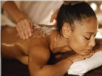 A woman receiving a back massage with salt or exfoliant applied to her shoulders, lying face down on a massage table.