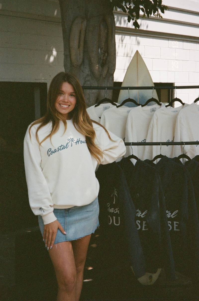 Megan, founder of Saltrock Studios, in a white Coastal House sweatshirt and blue skirt, standing by a rack of sweatshirts