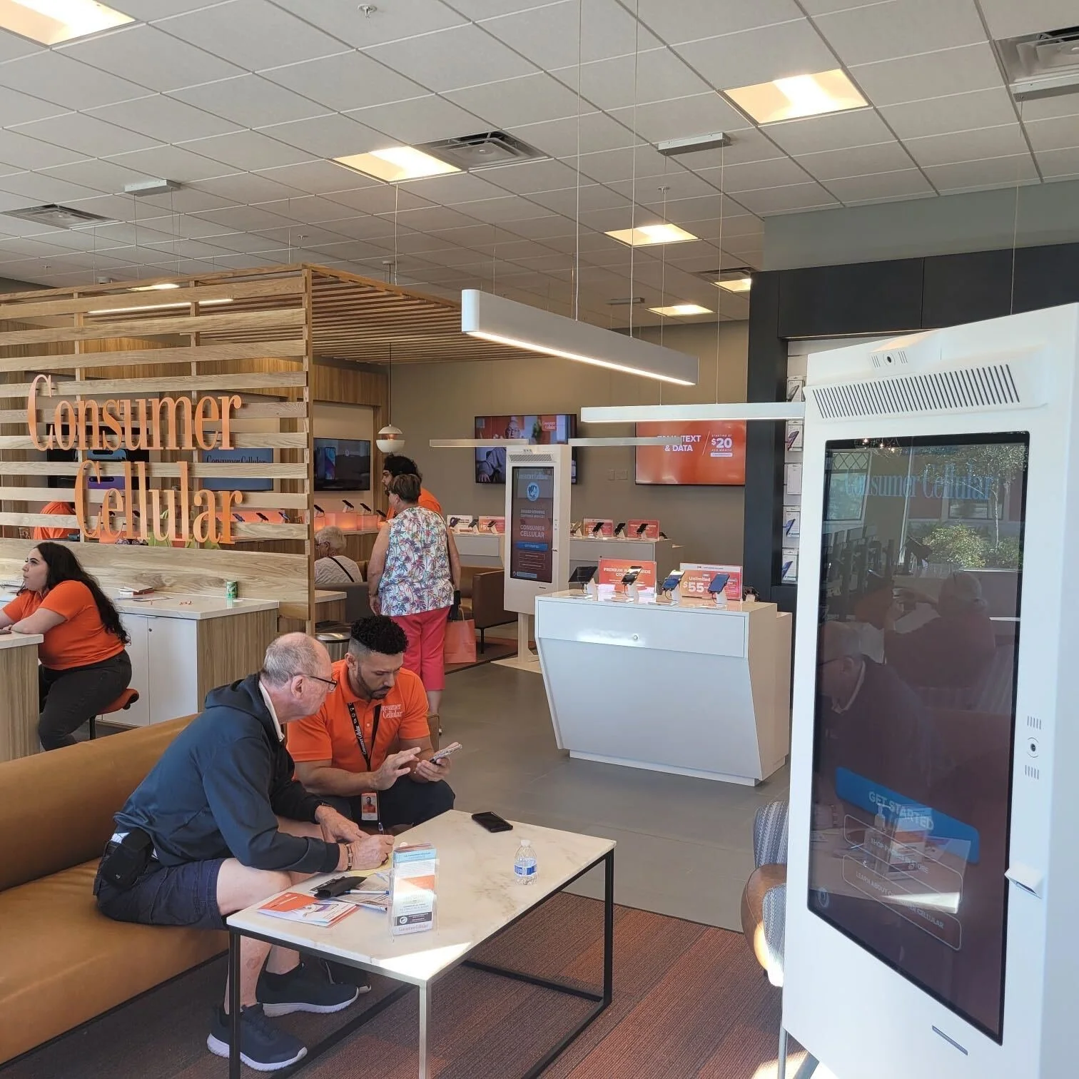 Inside an electronics store showing people shopping, sitting, and interacting, with a sign that reads 'Consumer Cellular' and several electronic devices on display.