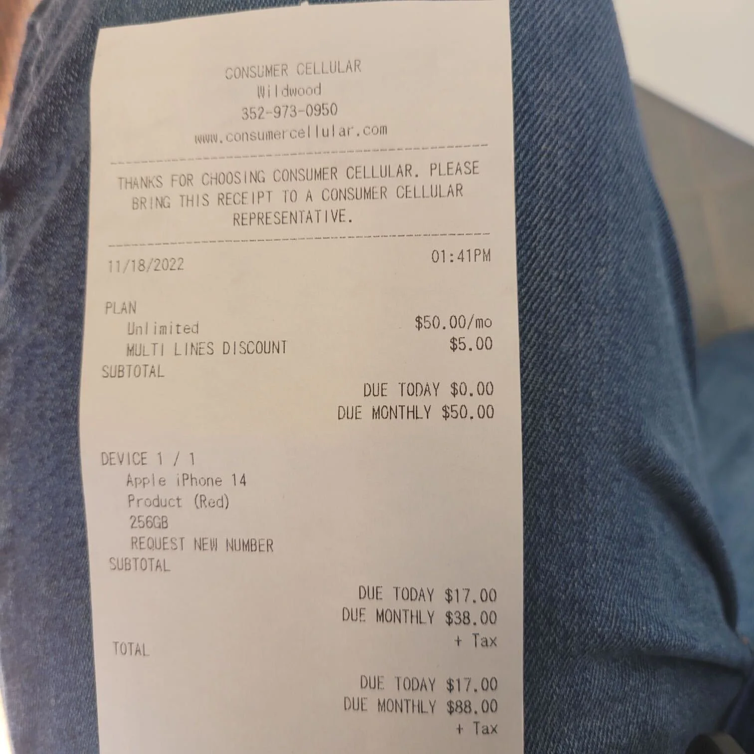 A printed receipt from Consumer Cellular showing details of a purchased Apple iPhone 14, including plan, prices, and billing information, resting on denim fabric.