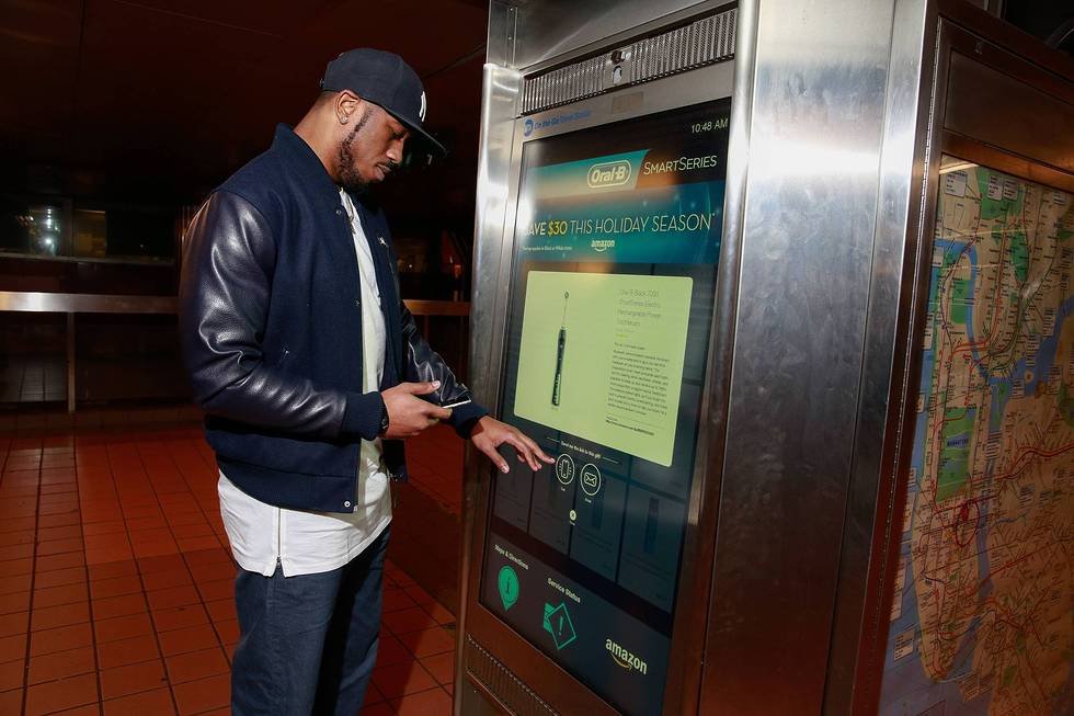 A man in a black jacket and baseball cap using a touchscreen kiosk at a transportation station, with a map on the wall to the right.