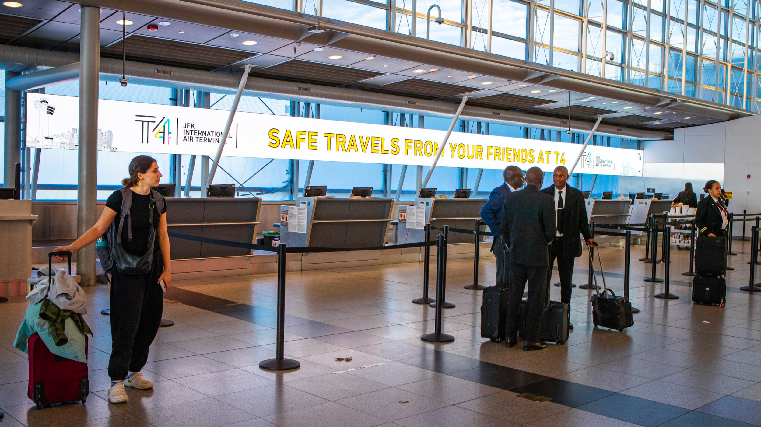 Passengers at JFK Airport's international terminal check-in counters with luggage, a woman with a rolling suitcase near the counter, and others chatting with airline staff, in a spacious, modern terminal with large windows.