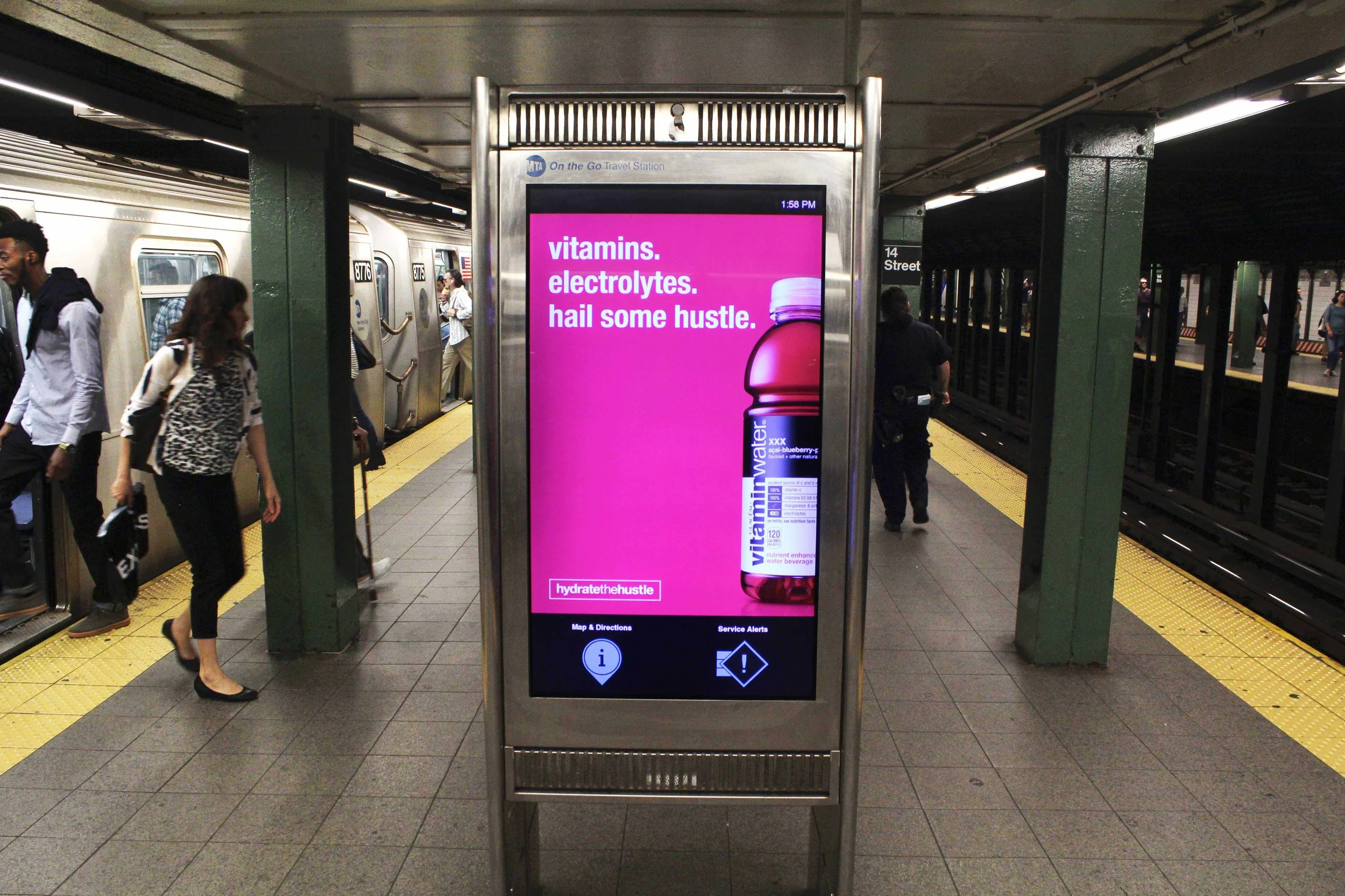 Digital advertisement on a subway platform promoting Vitamins water with the slogan 'vitamins. electrolytes. hail some hustle.' The ad features a pink background and an image of a pink water bottle.