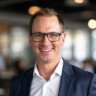 A smiling man wearing glasses, a white shirt, and a dark blazer in a professional setting.