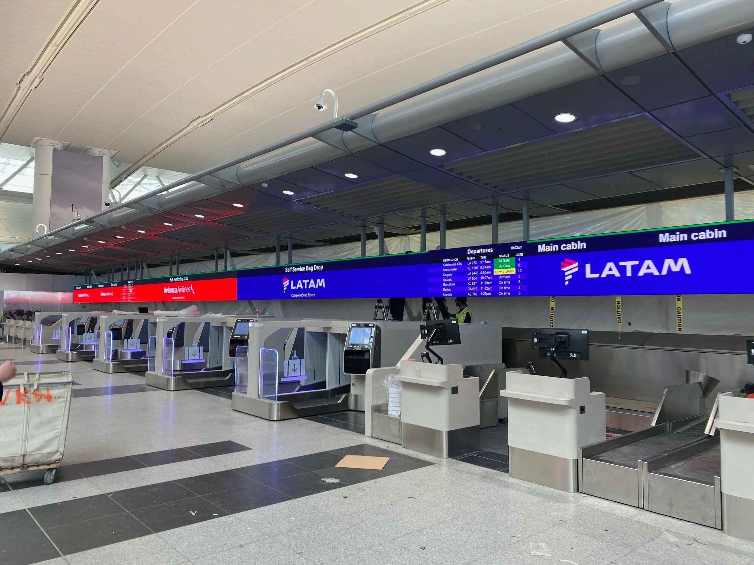 Empty check-in counters at the airport with electronic devices and luggage carts, digital departure information board overhead showing flight details.