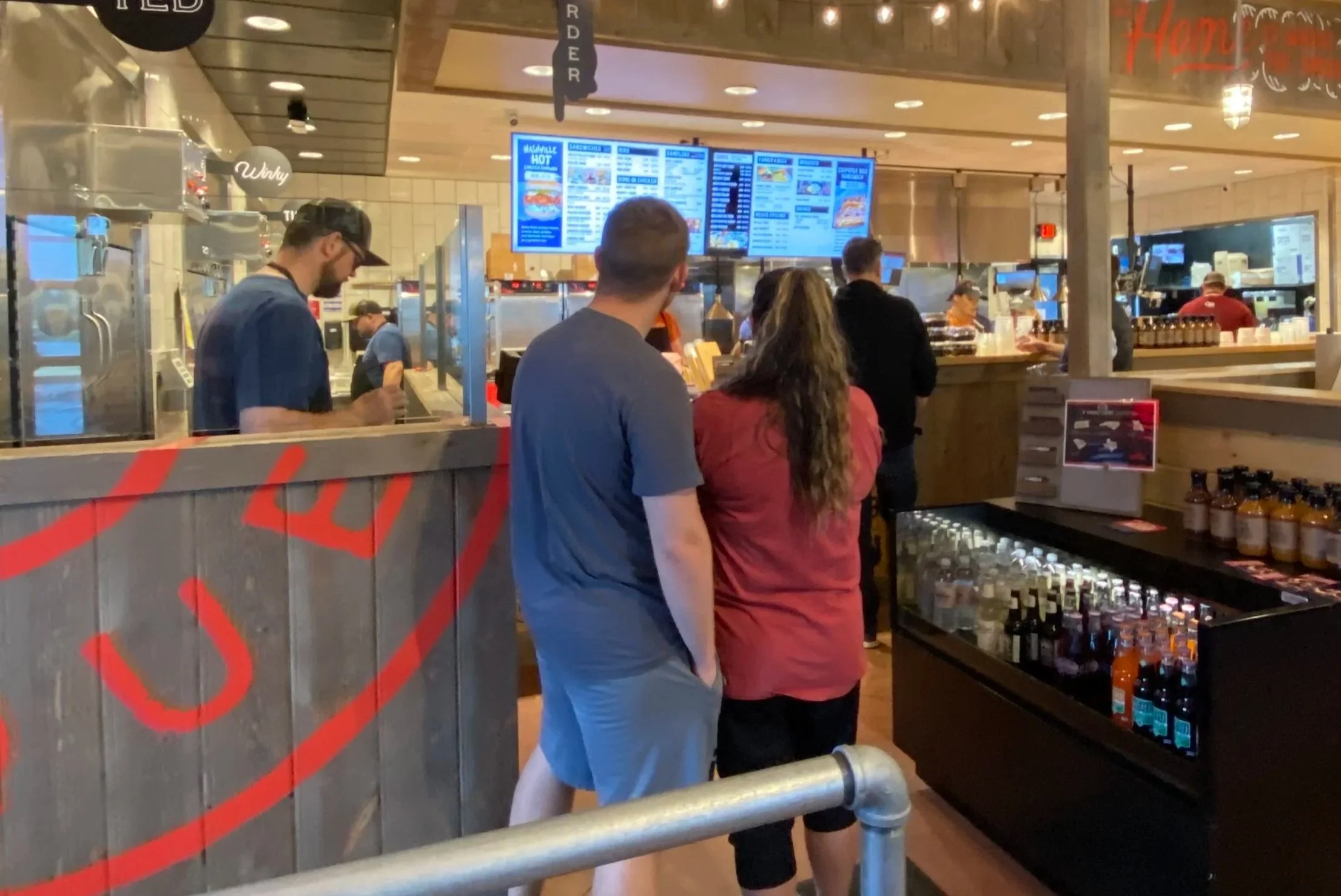 People ordering food at a fast-food restaurant counter with digital menu screens overhead.