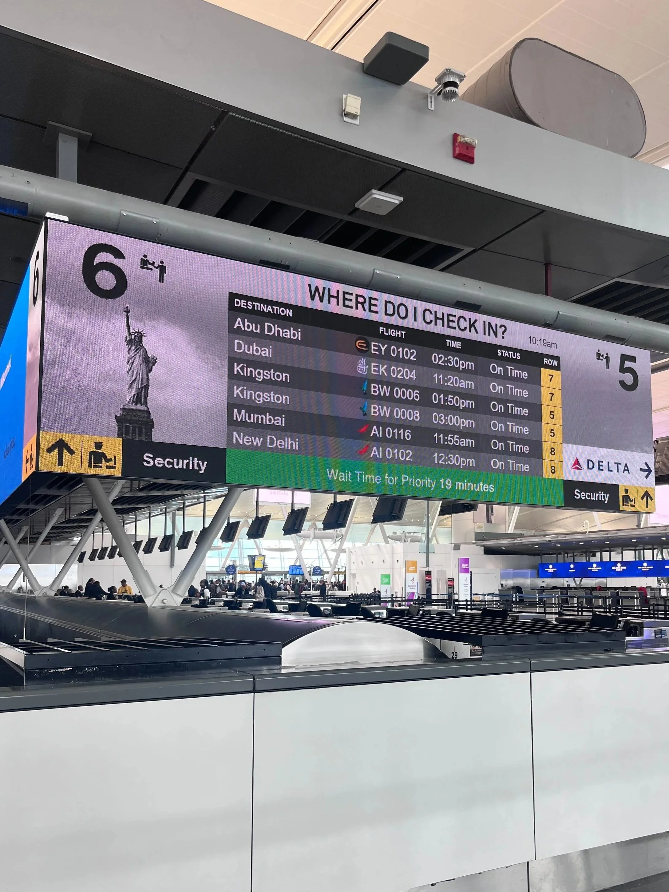 Airport baggage claim area with overhead electronic flight information display showing flights to Abu Dhabi, Dubai, Kingston, Mumbai, and New Delhi, with the time and status of each flight.