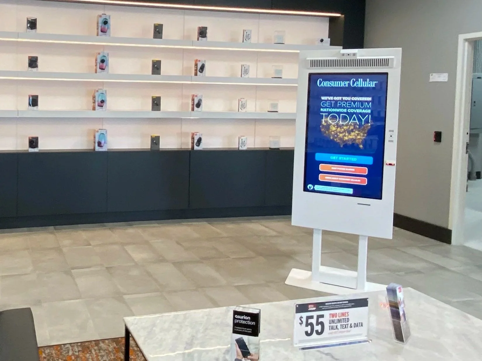 Inside a retail store with empty shelves displaying boxed mobile phones and a digital kiosk advertising a mobile plan. A table in the foreground has advertising signs for a mobile plan priced at $55 offering unlimited talk, text, and data.